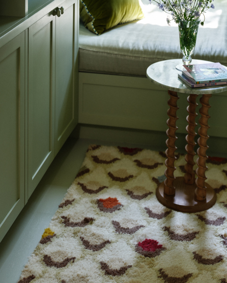 Bedroom corner with white dresser, bed with yellow pillow, patterned rug with brown crescents and colorful dots, and wooden side table with vase.