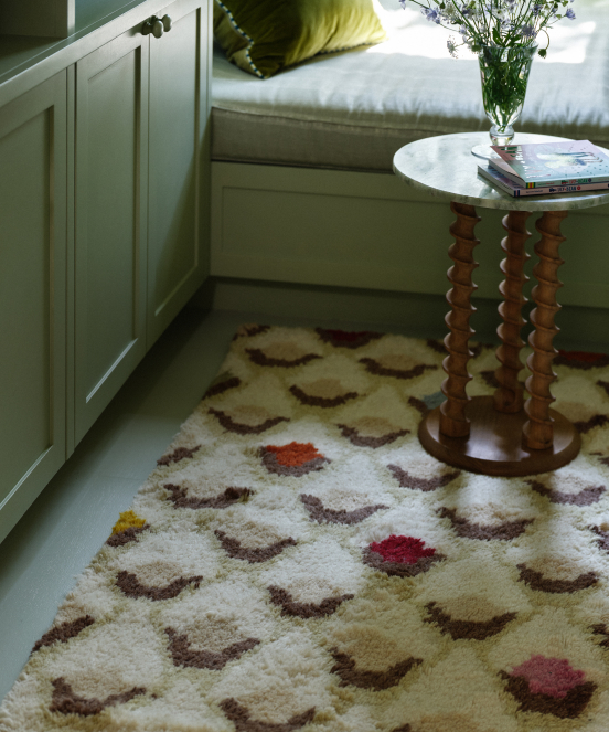 Cream shag rug with brown scalloped pattern and colorful dots beside a bed with white paneled base and round side table