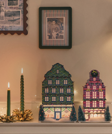 Two decorative miniature houses with lit windows displayed on a mantel with candles and small trees beneath framed photos.
