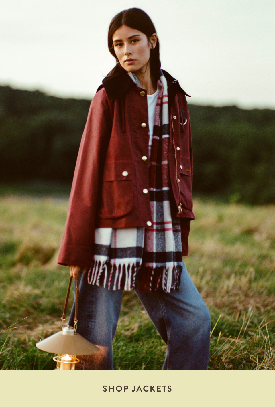 Woman in burgundy jacket and plaid scarf standing in countryside field holding lantern with text SHOP JACKETS below