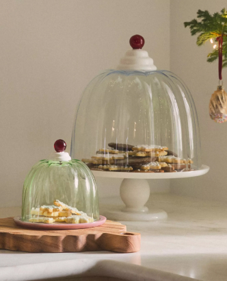 Glass cake domes with red knob finials covering cookies on white and wooden cake stands in a holiday setting.
