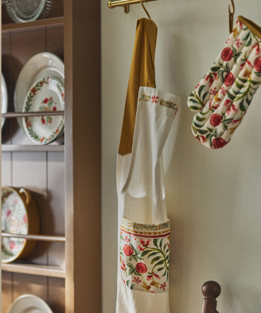 White apron with red and green floral embroidery hanging on wooden hanger, with matching plates displayed in cabinet behind