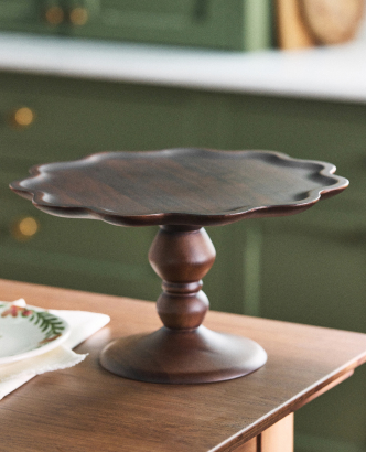 Bronze-colored metal cake stand with scalloped edge and pedestal base on wooden table against green wall background.