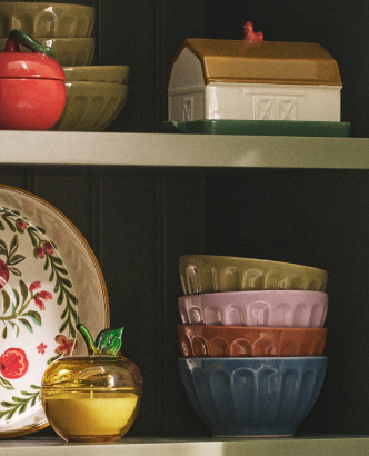 Shelves displaying vintage kitchenware including colorful bowls, a red apple container, decorative plates, and a barn-shaped container.