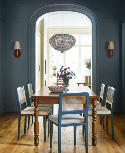 Elegant dining room with dark teal walls, wooden table with caned chairs, decorative pendant light, and arched doorway leading to bright space.