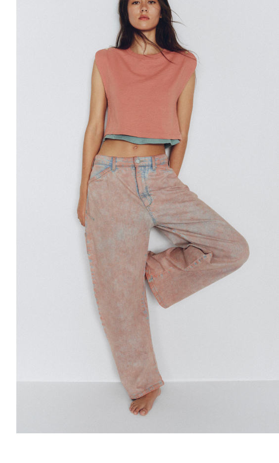Woman wearing coral sleeveless crop top with teal trim and beige wide-leg jeans against white background