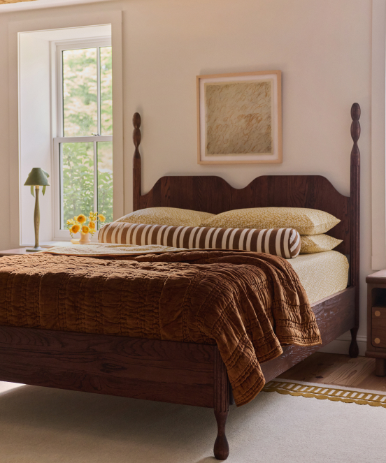 Cozy bedroom with dark wood four-poster bed, brown quilted bedding, striped pillows, framed art, window with greenery view, and lamp.