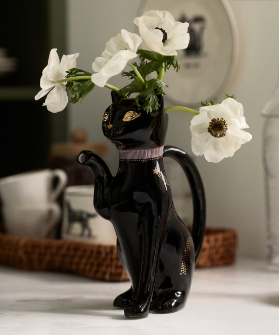 Black cat-shaped vase with white anemone flowers on a kitchen counter with woven basket and mugs in background