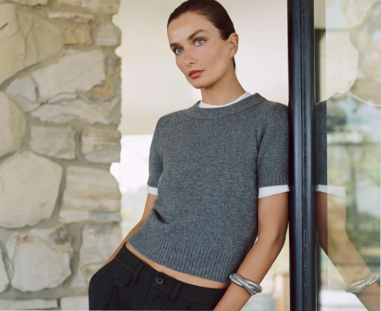 Woman wearing gray short-sleeve knit sweater with white trim, standing by stone wall and black-framed window