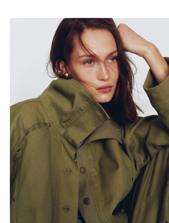 Woman with brown hair wearing an olive green button-up jacket, hand in hair, looking at camera against white background.