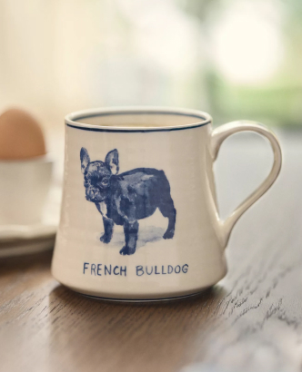 White ceramic mug with blue French Bulldog illustration and text "FRENCH BULLDOG" on wooden surface with blurred background.