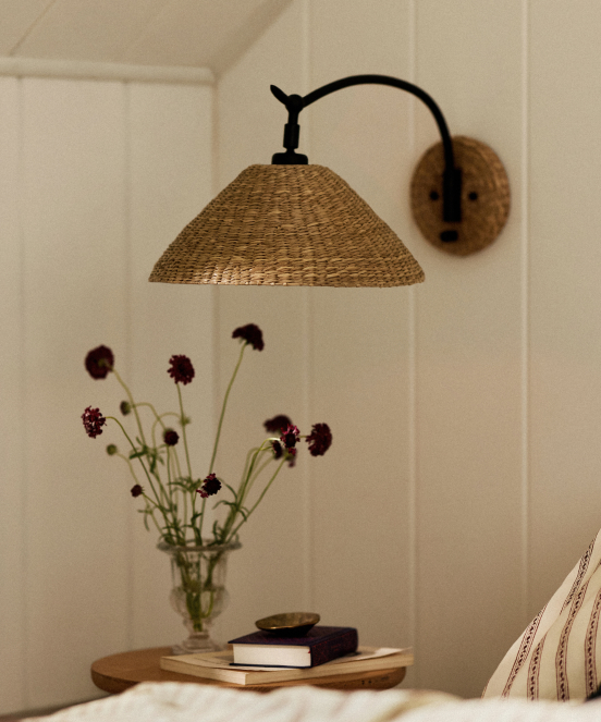 Woven rattan wall sconce with curved arm above a bedside table with purple flowers in a glass vase and stacked books.