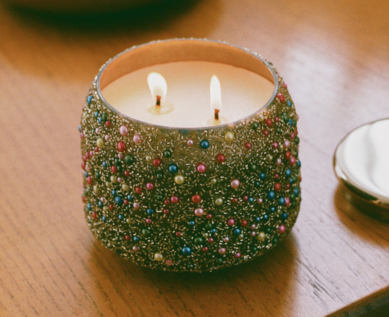 Decorative candle with two lit wicks in a green glitter and multicolored gem-studded container on a wooden surface.