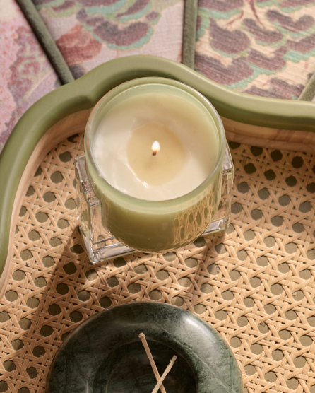Lit candle in green glass jar on woven rattan tray with decorative items and patterned background