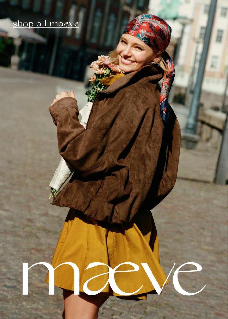 Smiling woman in colorful headscarf, brown jacket, and yellow skirt holding flowers outdoors. Text reads shop all maeve.