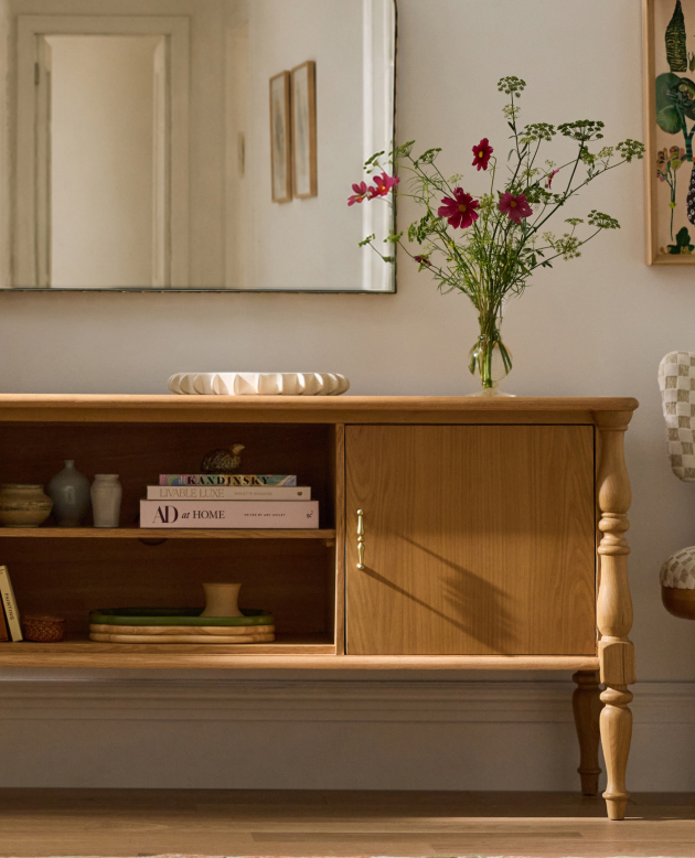 Wooden console table with open shelving displaying books and decor, topped with a vase of pink cosmos flowers and mirror above.