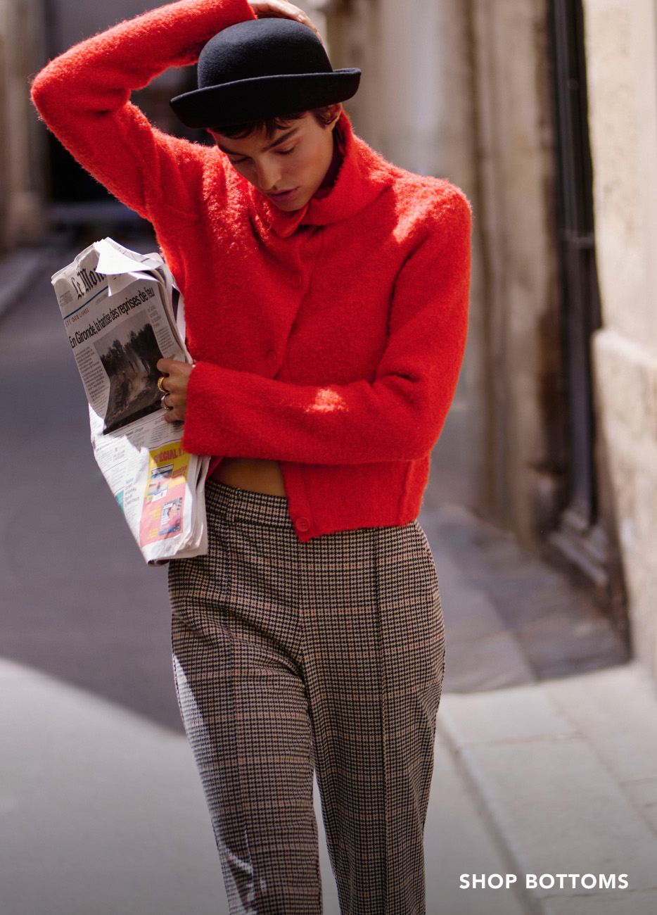 Woman in bright red sweater and black bowler hat holding newspaper, wearing checkered pants. Text reads SHOP BOTTOMS.