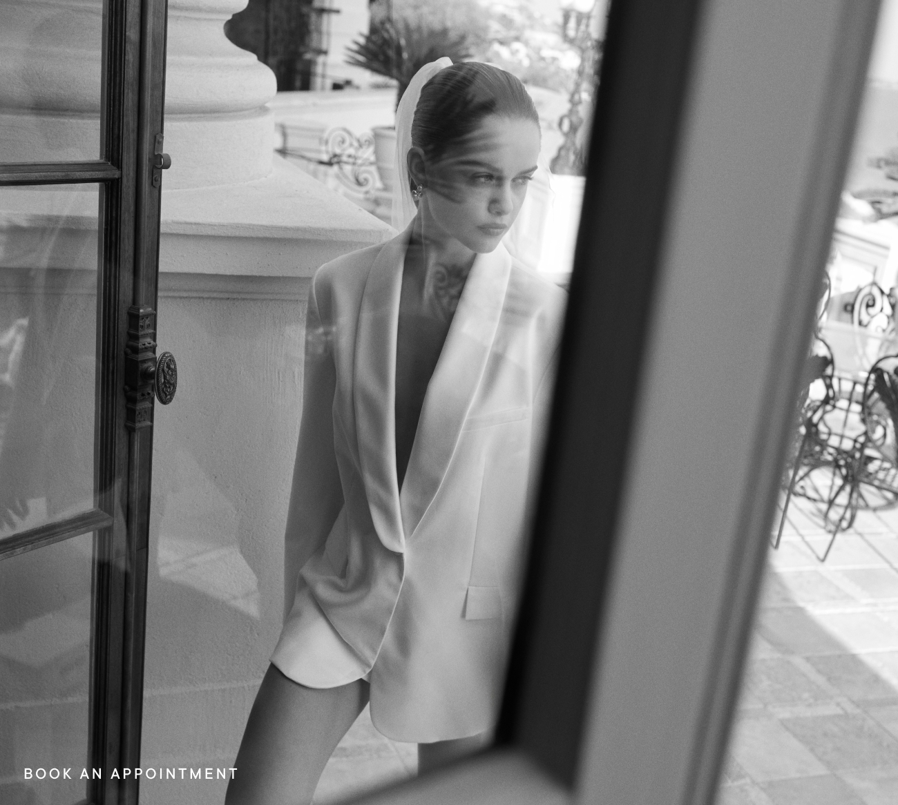 Black and white photo of woman in blazer standing by doorway with ornate ironwork visible outside