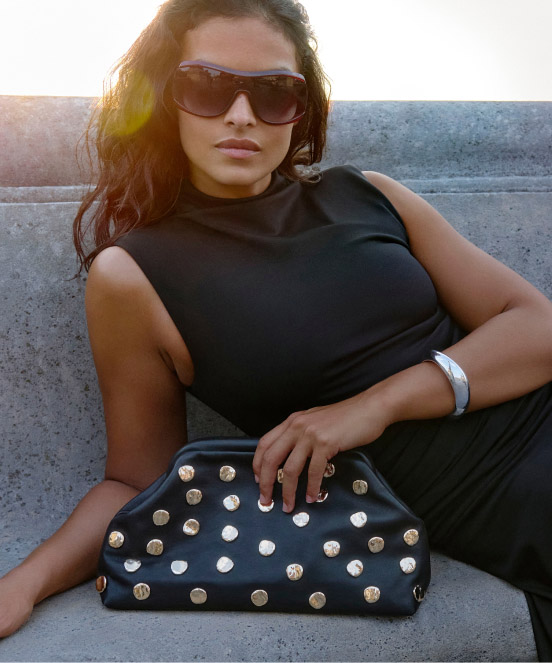 Woman in black sleeveless top and sunglasses sitting on concrete steps, holding a black studded handbag with gold hardware.