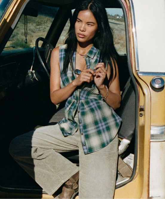 Woman with long dark hair wearing green plaid sleeveless shirt and beige pants sitting in doorway of yellow vintage truck