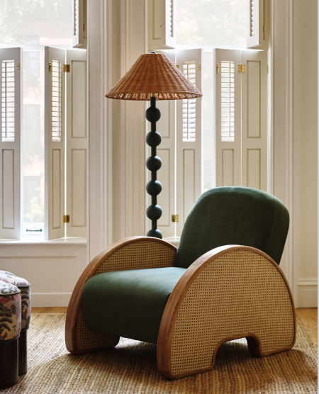Green velvet armchair with woven rattan sides next to a tall black sculptural floor lamp with wicker shade in a bright room.