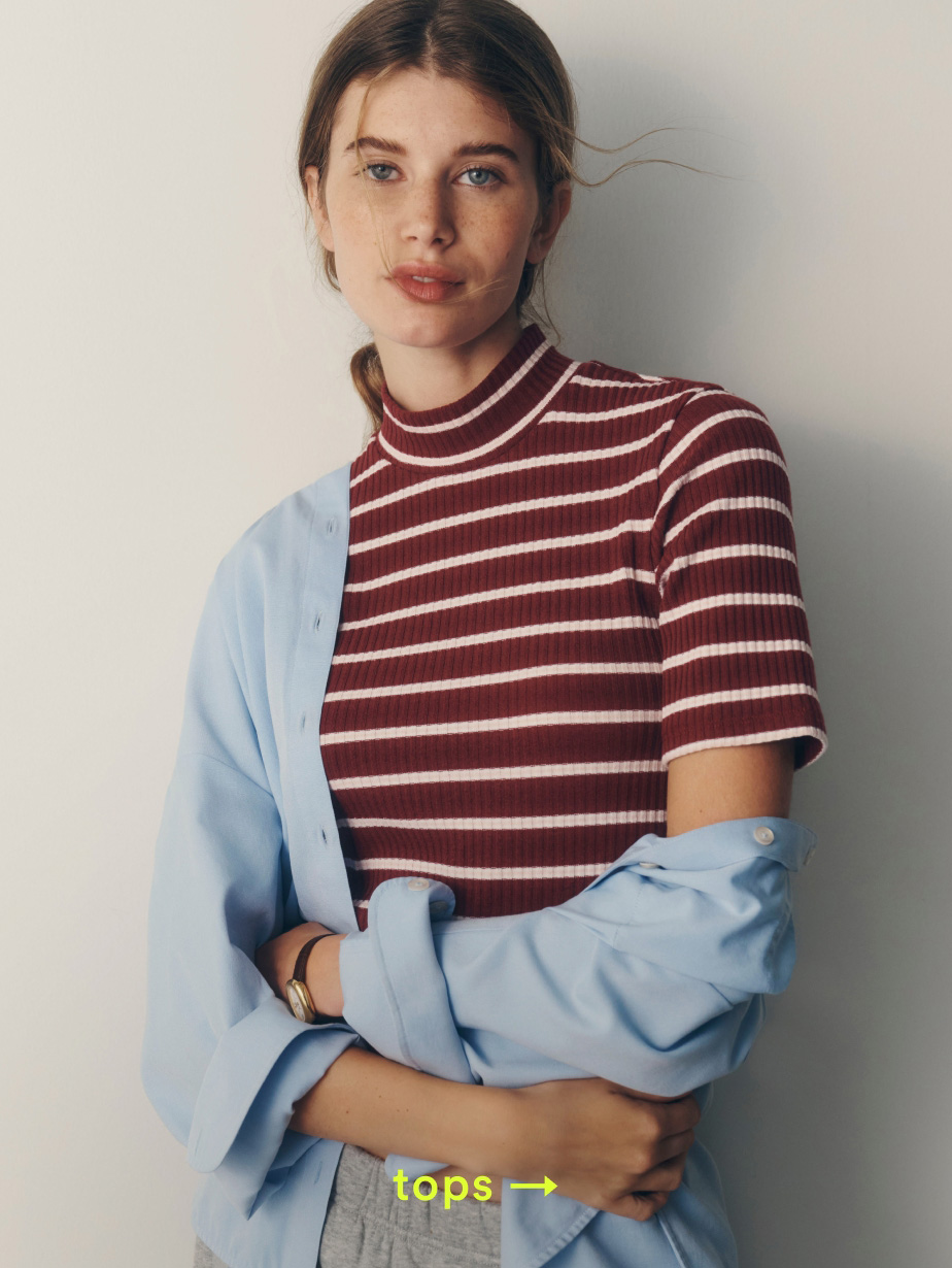 Woman wearing burgundy and white striped ribbed top with light blue shirt draped over shoulders. Yellow text reads "tops →"