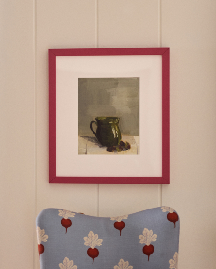 Framed painting of a dark green pitcher with white matting and red frame, hung on white paneled wall above blue fabric with beet pattern.