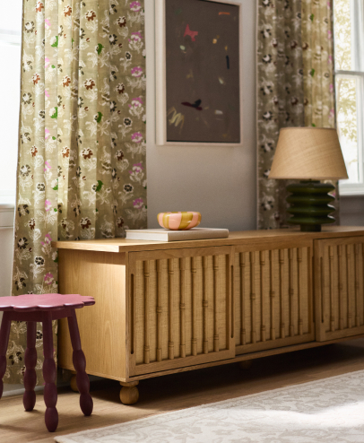 Living room with wooden sideboard, floral curtains, beige lamp, framed artwork, purple stool, and decorative bowl on books.