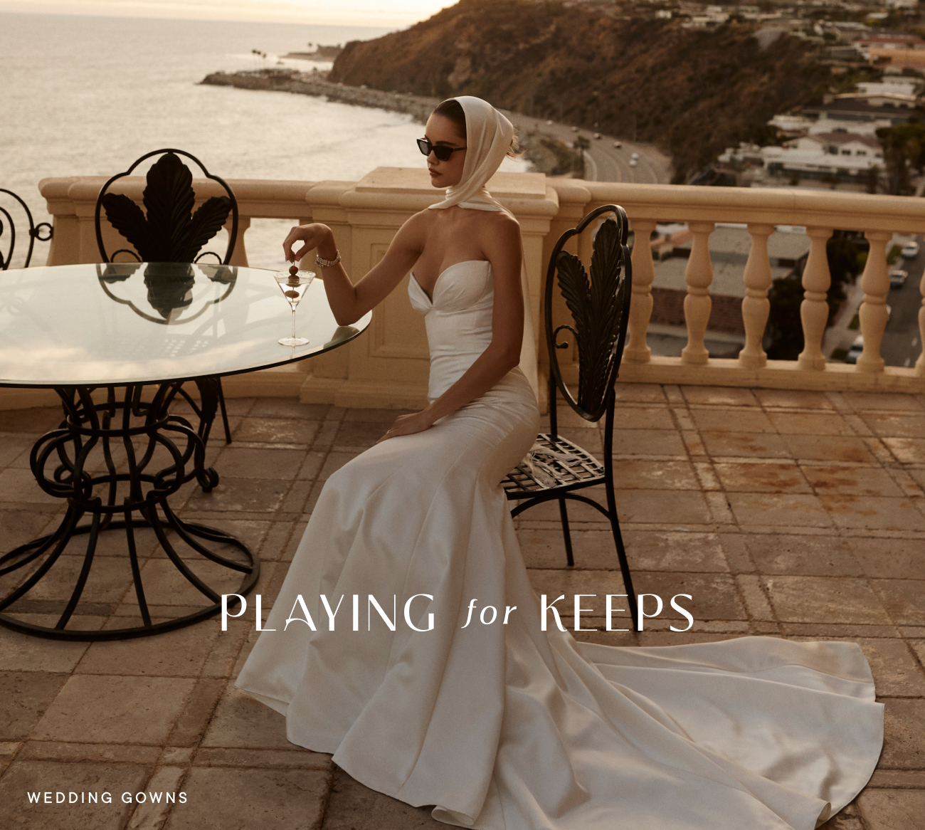 Woman in white strapless gown and headscarf sits at coastal terrace table with martini. Text reads PLAYING for KEEPS.