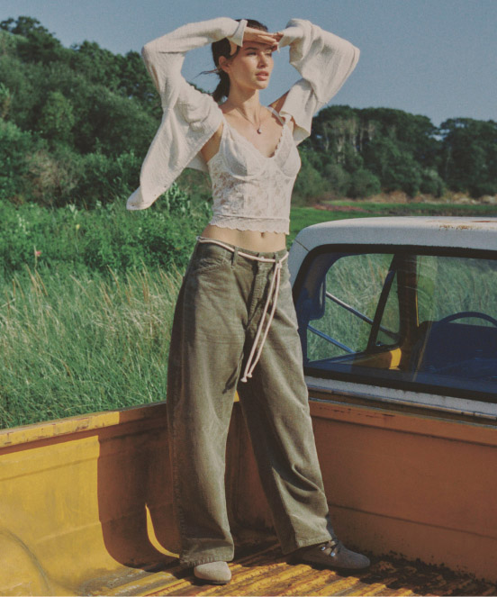 Woman in white cropped blouse and olive wide-leg pants stands by orange vintage car door in grassy field with trees.