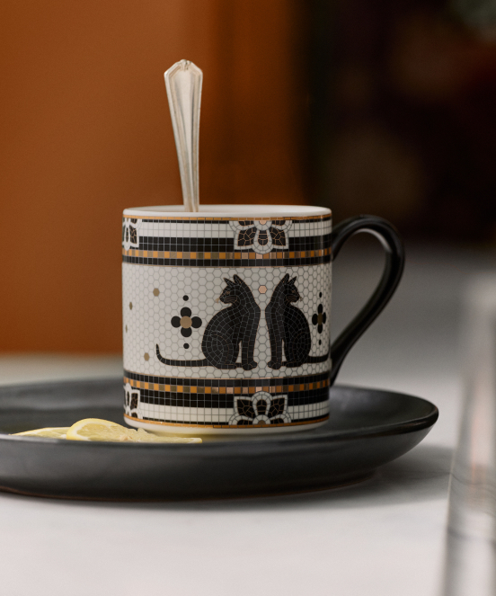 Decorative mug with black cat silhouettes and paw prints on a black saucer with a white spoon inside against a blurred background.