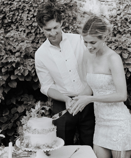 Black and white photo of a couple cutting their wedding cake together outdoors with ivy-covered wall in background
