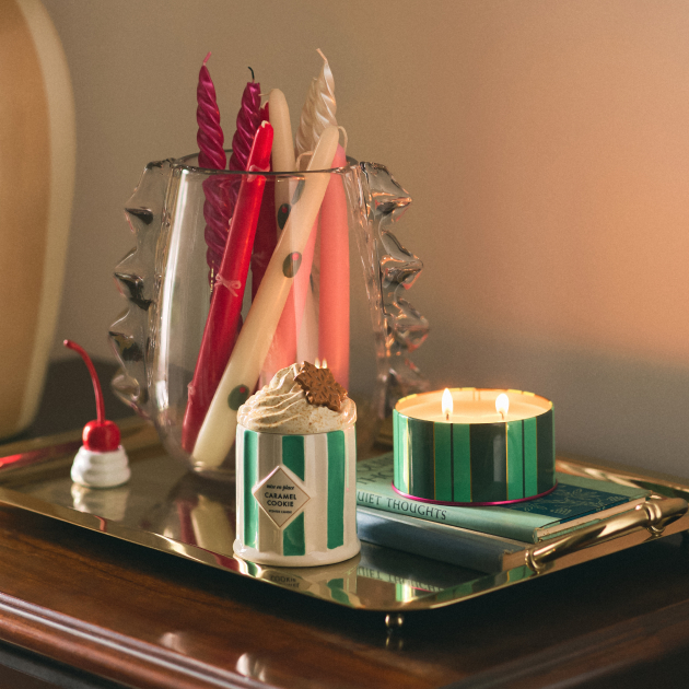 Decorative candles in various shapes and colors displayed on a gold tray with a lit green-striped candle and cherry ornament.