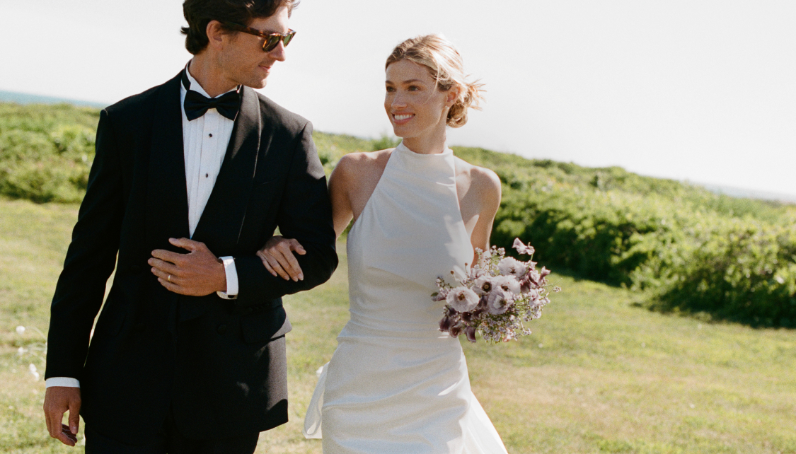 Smiling bride in white halter gown with bouquet walks arm-in-arm with groom in black tuxedo on grassy coastal hillside.