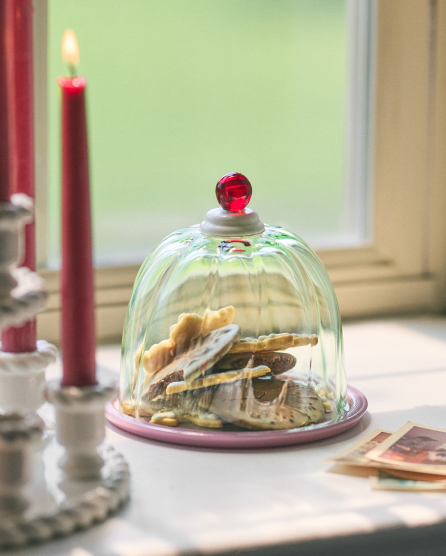 Glass dome with red knob covering cookies on a plate, with red taper candles in holders nearby on a white surface.