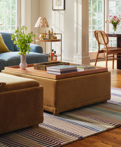 Bright living room with wooden storage ottoman coffee table, blue sofa, striped rug, and large windows with natural light.
