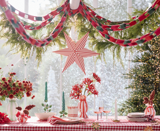 Festive holiday table setting with red and white checkered tablecloth, striped star decoration, garland, and red flowers in vase.