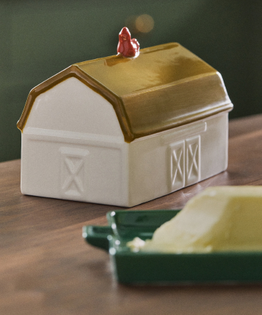 White ceramic barn-shaped container with gold lid and small red rooster figure on top, sitting on a wooden surface.
