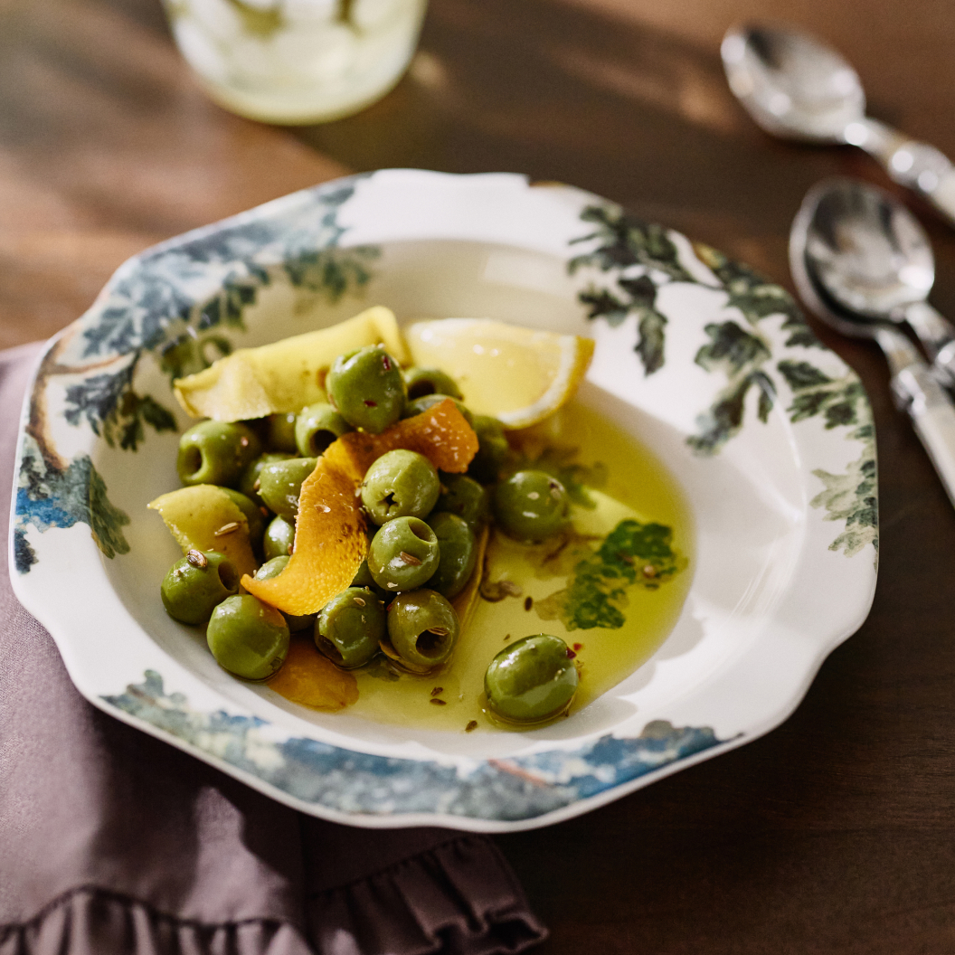 Green olives with lemon slices and orange peel in olive oil on a decorative white plate with green leaf pattern.