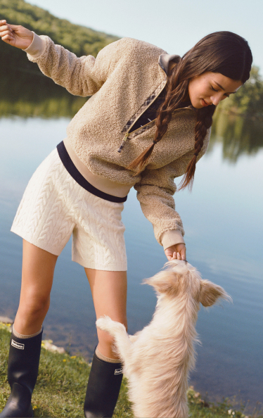 Woman in beige fleece jacket and cream shorts playing with a fluffy white dog by a lake with hills in the background.