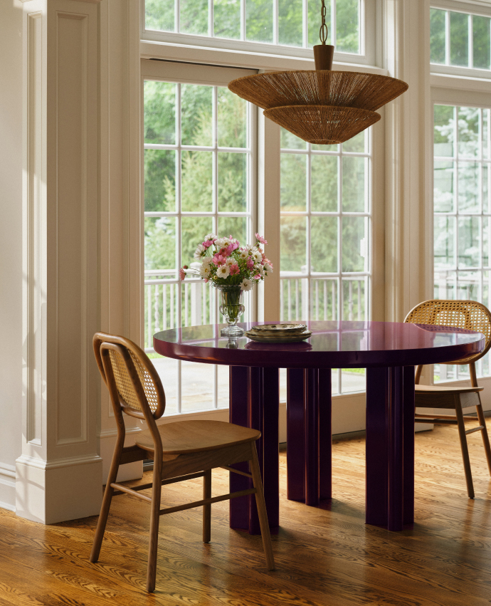 Round dark wood dining table with cane-back chairs, pink flower centerpiece, and woven pendant light in bright room with large windows.