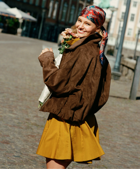 Smiling woman in colorful headscarf, brown jacket, and yellow skirt holding flowers on a city street.
