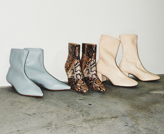 Three pairs of ankle boots in light blue, snakeskin print, and cream displayed on concrete floor against white wall.