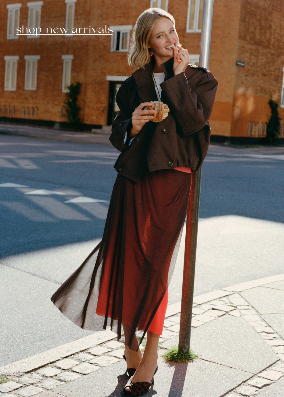 Woman in brown blazer and red skirt eating pastry on city street with text shop new arrivals in upper left corner