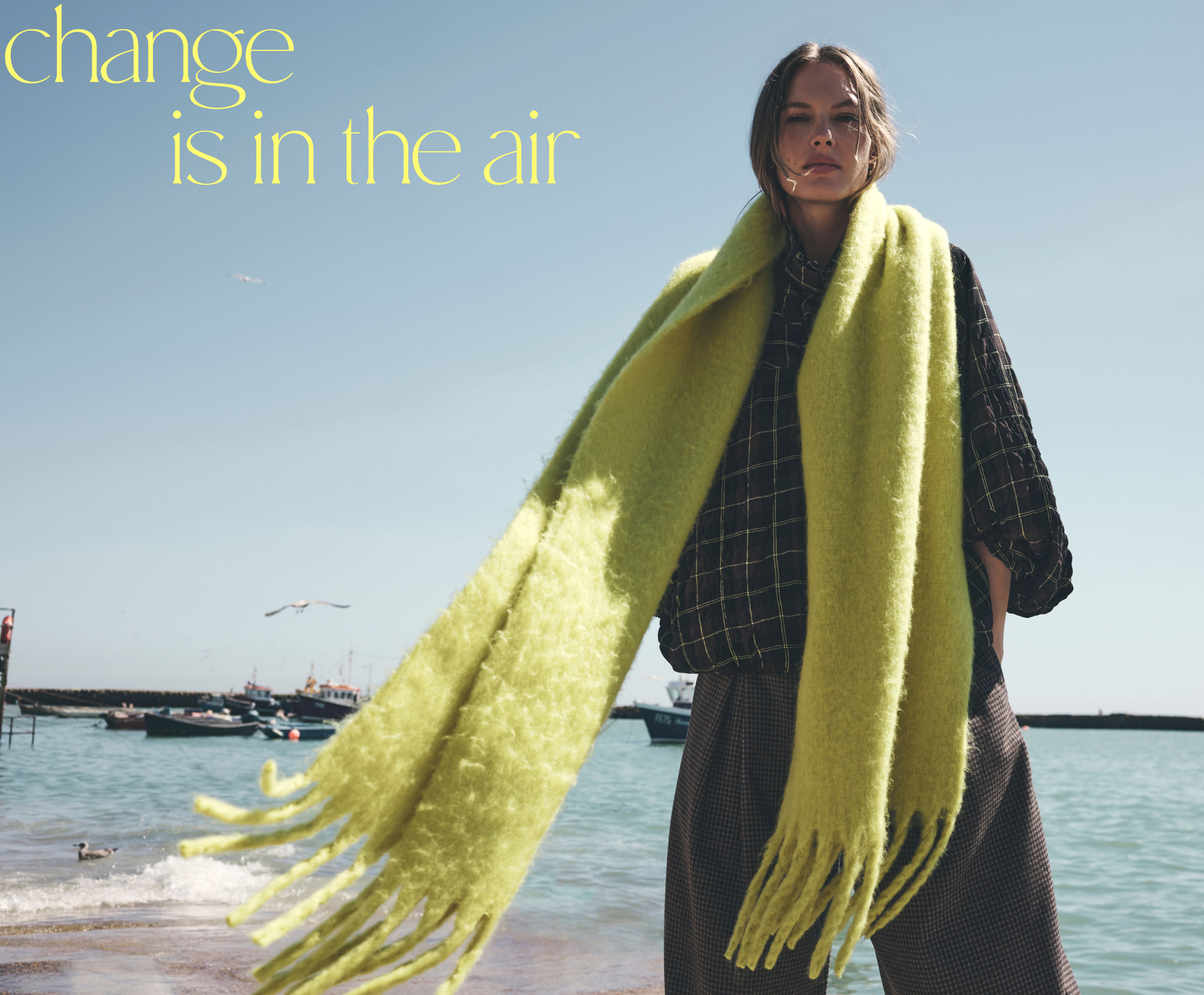 Woman in black checkered outfit with bright yellow-green scarf at beach with text "change is in the air" on blue sky.