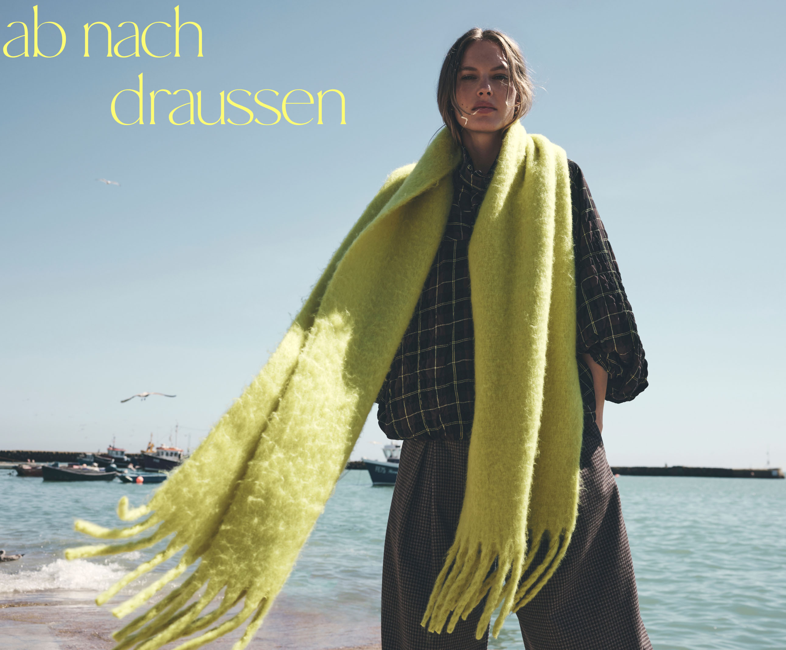 Woman in black checkered outfit with bright yellow-green scarf at beach with text "change is in the air" on blue sky.