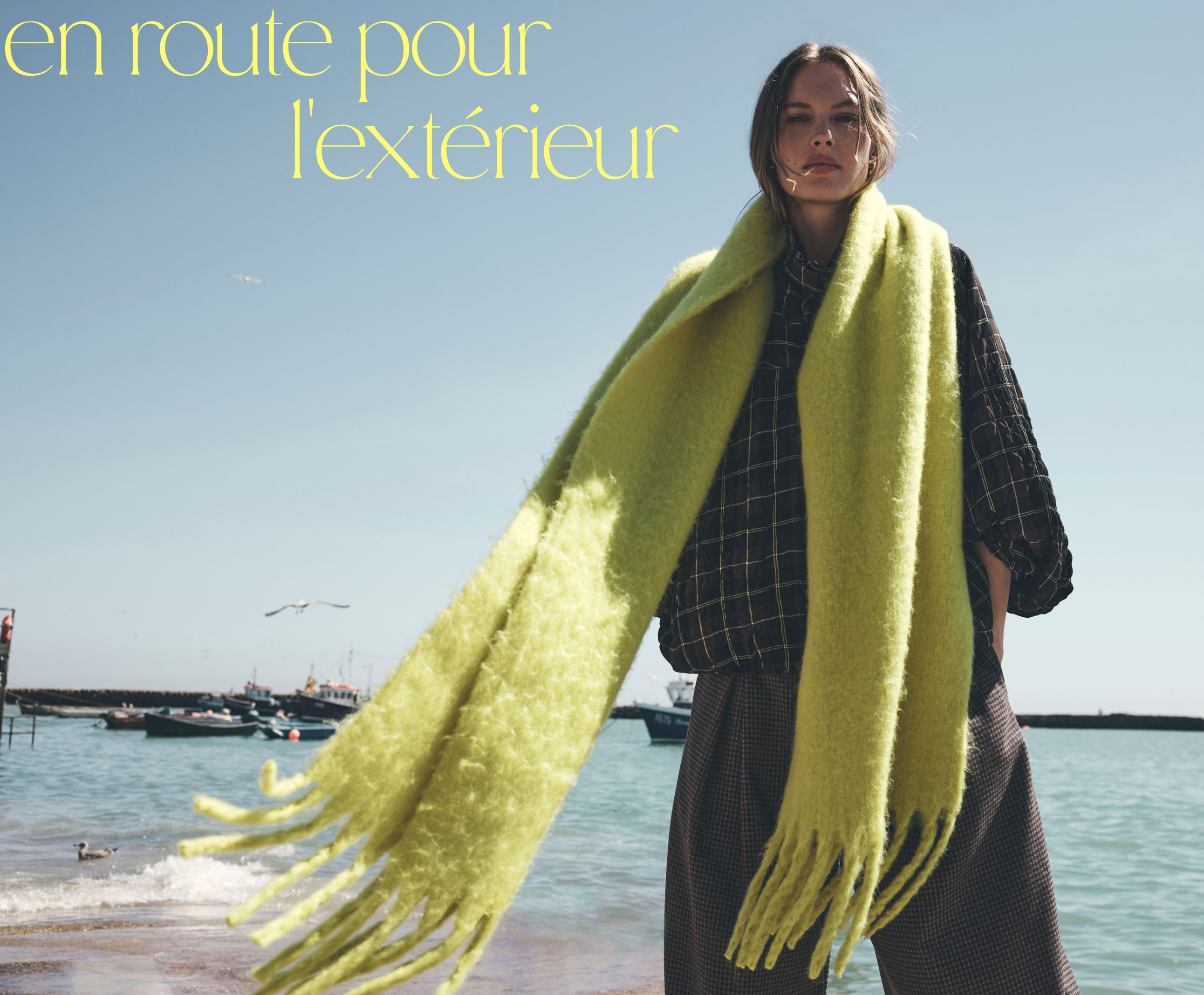 Woman in black checkered outfit with bright yellow-green scarf at beach with text "change is in the air" on blue sky.