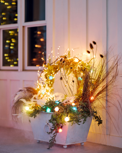 White planter with dried grasses, evergreen branches, and colorful string lights on a porch with windows in background.