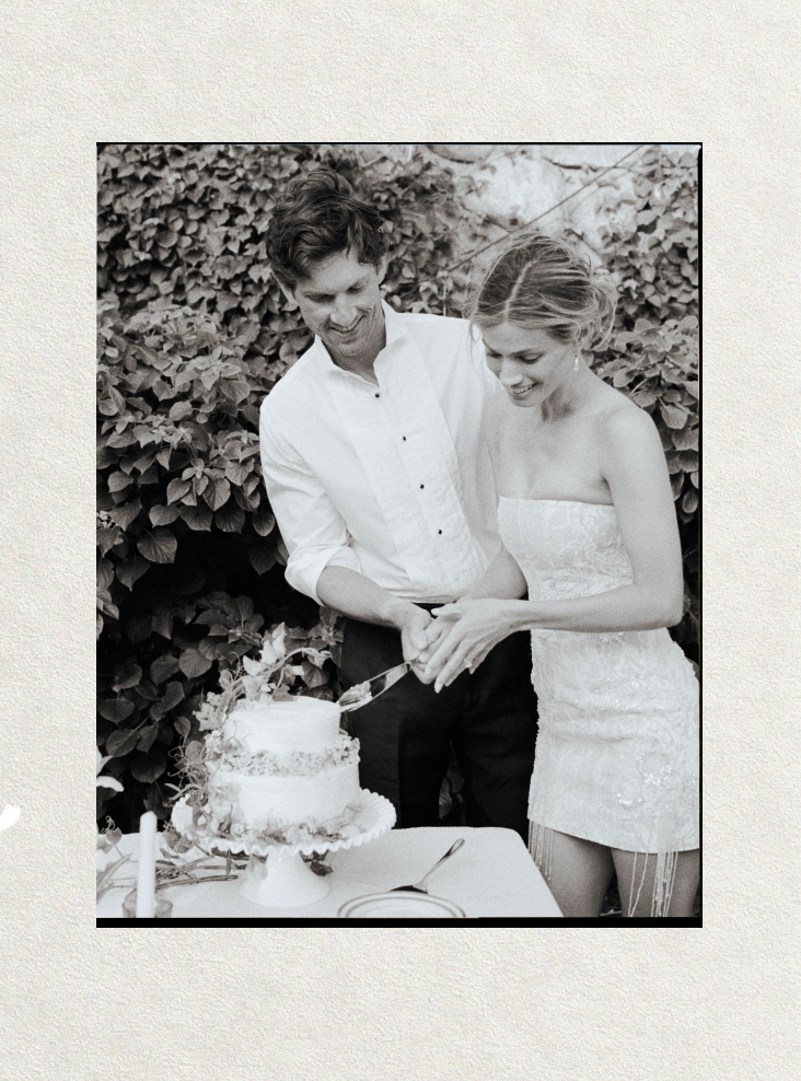 Black and white photo of bride and groom cutting their white wedding cake together outdoors with ivy backdrop