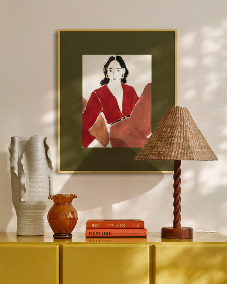 Framed portrait of woman in red jacket above yellow console table with wicker lamp, orange vase, books, and white sculpture